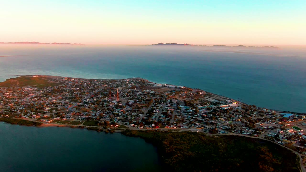 Aerial dusk view over San Carlos, Baja California Sur, with a dark lagoon in the foreground, a dense coastal town on a peninsula, open ocean beyond, distant islands on the horizon, and no visible people.