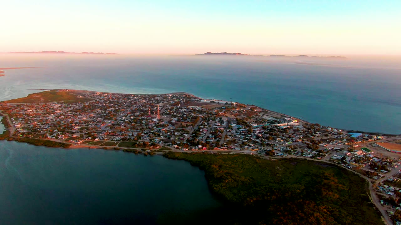 Aerial view over San Carlos, Baja California Sur, with clustered low buildings, a tall communications structure near the town center, lagoon water below, open ocean behind, and no visible people.