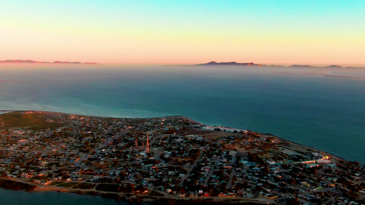 Aerial dusk view over San Carlos, Baja California Sur, with town blocks in the foreground, blue ocean beyond, distant islands on the horizon, and no visible people.