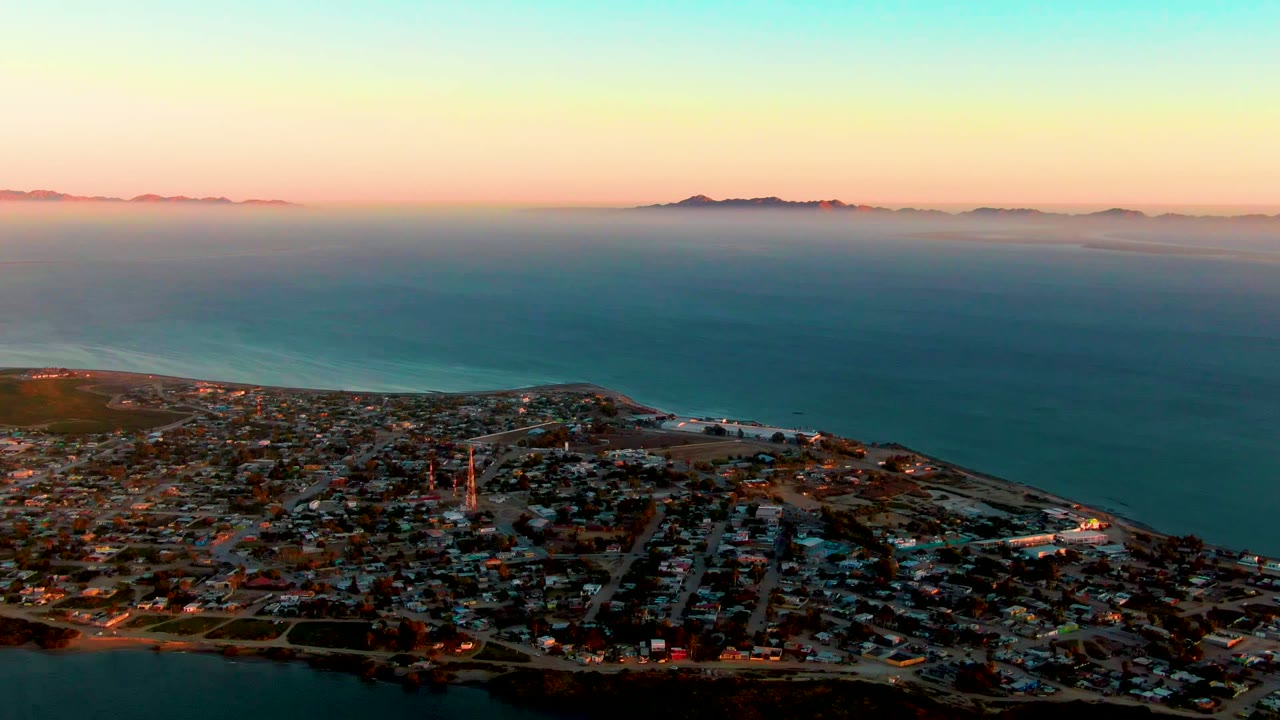 Wide aerial frame over San Carlos, Baja California Sur, with the town at the lower edge, open blue ocean across the center, hazy islands on the horizon, and no visible people.