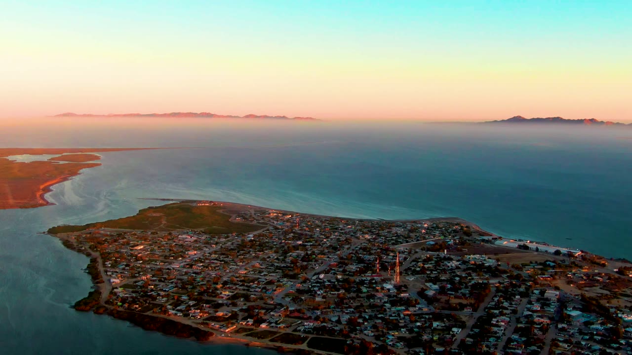 Aerial view over San Carlos, Baja California Sur with town rooftops below, calm sea beyond, distant mountains on the horizon, pink evening sky, and no visible people.