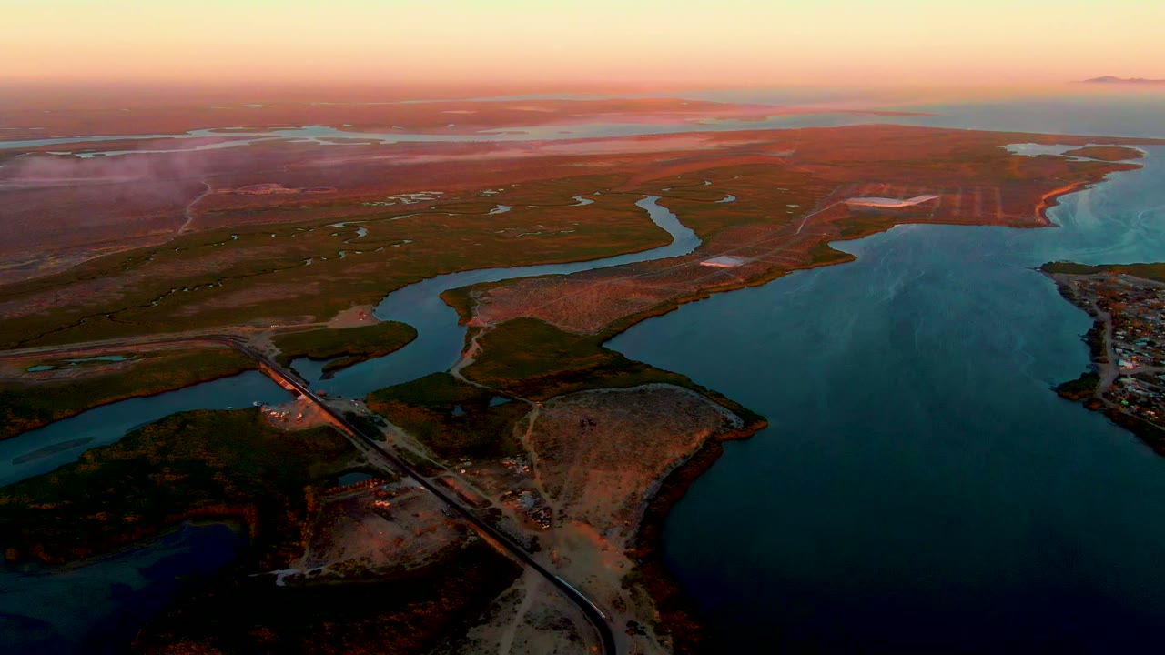 Aerial sunset view over San Carlos, Baja California Sur with a bridge in the lower left, blue water channels, reddish terrain, orange sky, and no visible people.