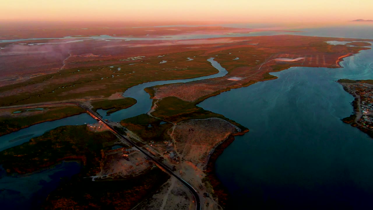 High aerial view in San Carlos, Baja California Sur with the bridge lower left, dark shoreline patches, winding water, reddish flats, soft orange horizon, and no visible people.