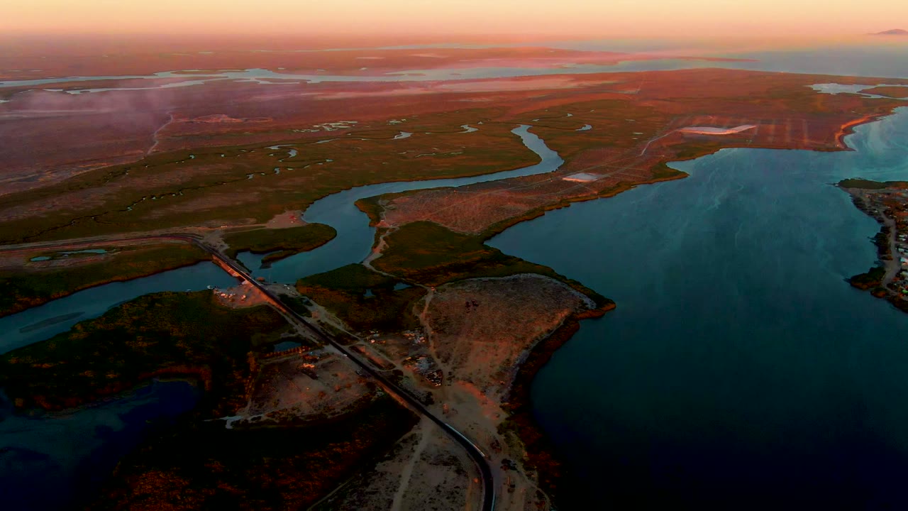 Wide drone view over San Carlos, Baja California Sur showing a blue water inlet, bridge and road at lower left, open brown terrain, low sunset sky, and no visible people.