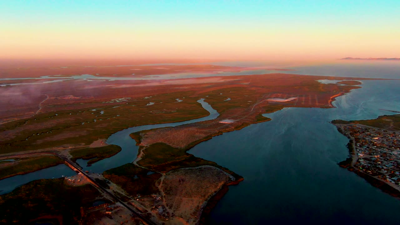 Aerial dusk view over San Carlos, Baja California Sur, with a bridge crossing water in the lower left, a shoreline settlement at right, broad blue inlet water, reddish land, orange sky, and no visible people.