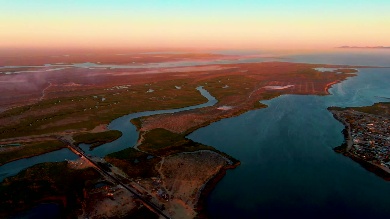 Wide aerial frame over San Carlos, Baja California Sur, with the water channel curving toward the center, bridge and road at the bottom, settlement on the right bank, flat land beyond, and no visible people.