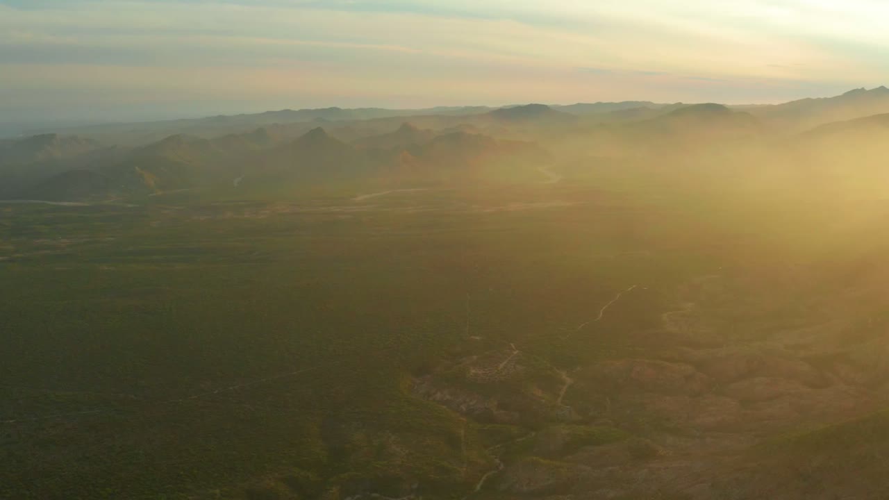 Aerial view over Cabo Pulmo, Baja California Sur with a green and brown valley, distant mountain ridges, soft clouds, and no visible people.