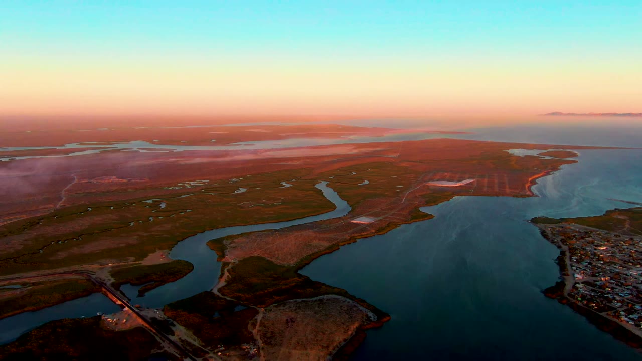 Aerial dusk frame over San Carlos, Baja California Sur, showing a winding coastal waterway, open reddish land, distant horizon water, and a small settlement at the lower right with no visible people.