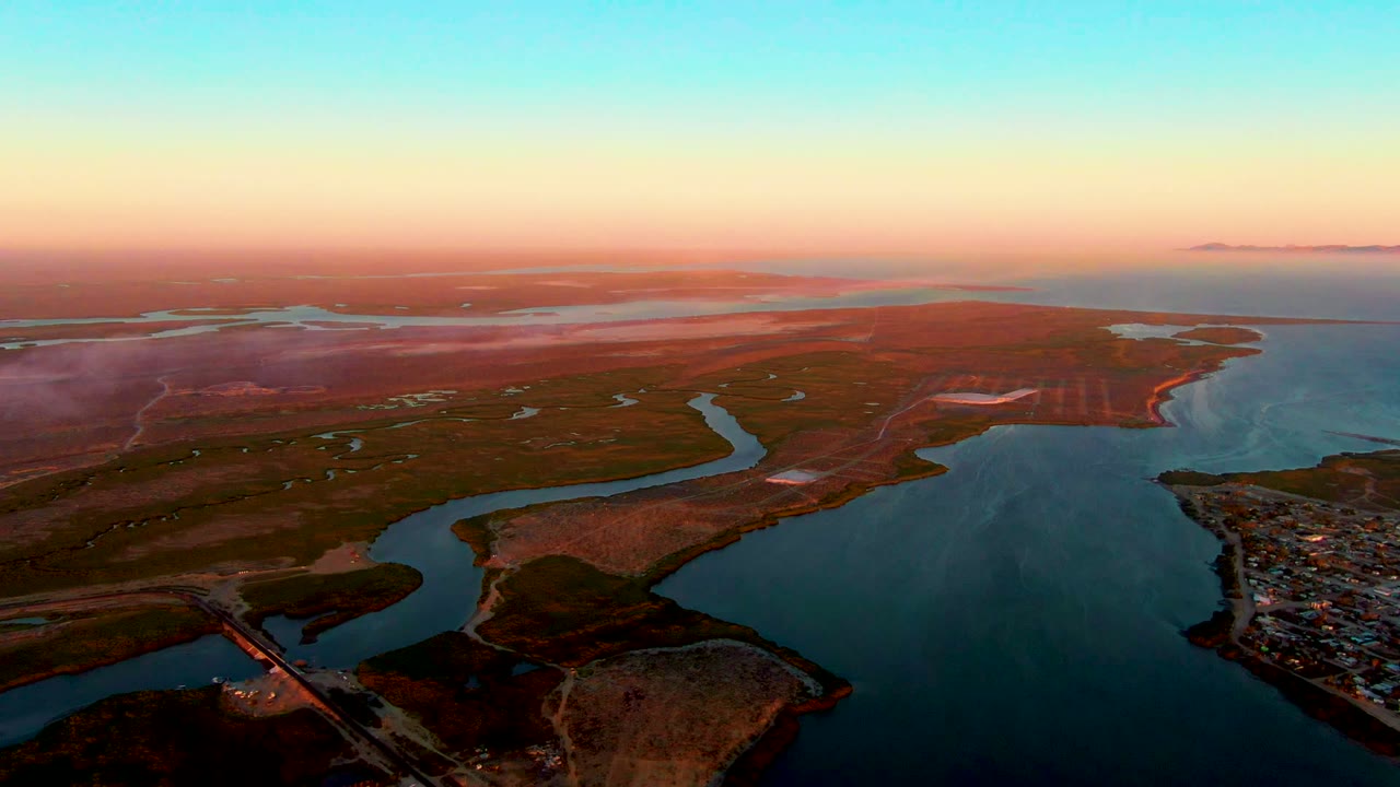 Elevated coastal waterway view with reddish land, blue water, and orange dusk horizon.