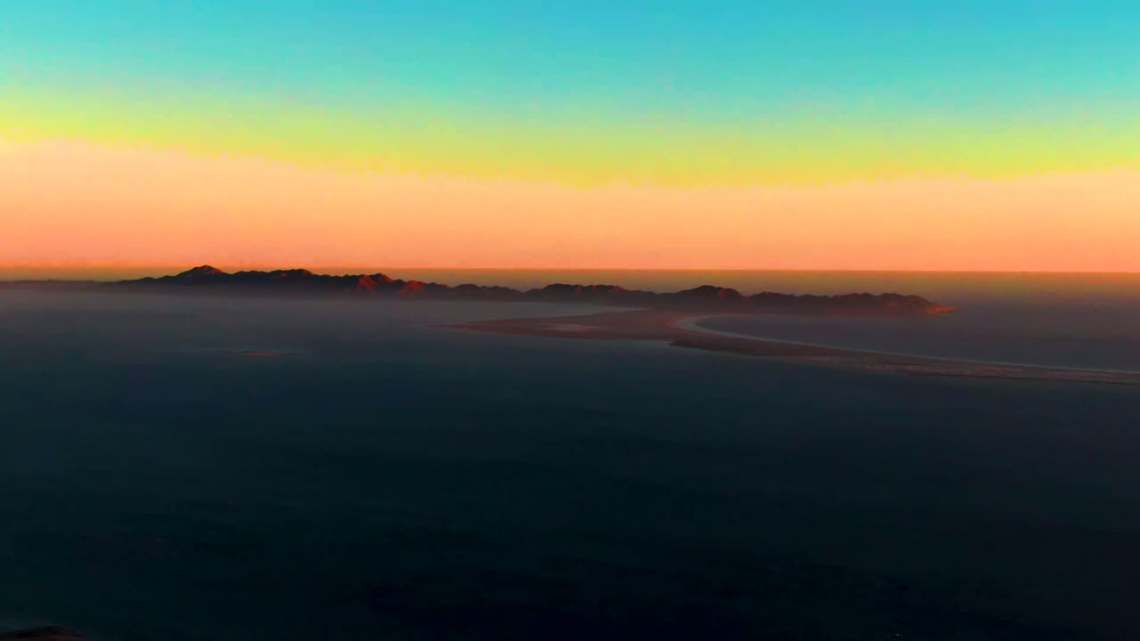 Wide aerial view over San Carlos, Baja California Sur at sunset, with dark sea below a rainbow-like sky band, distant mountain silhouettes, and no visible people.