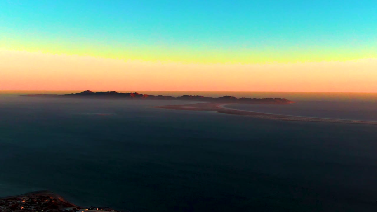 Aerial view of San Carlos at dusk with a low foreground town, small lights, bay water, distant mountains, and a fading orange-blue sky with no visible people.