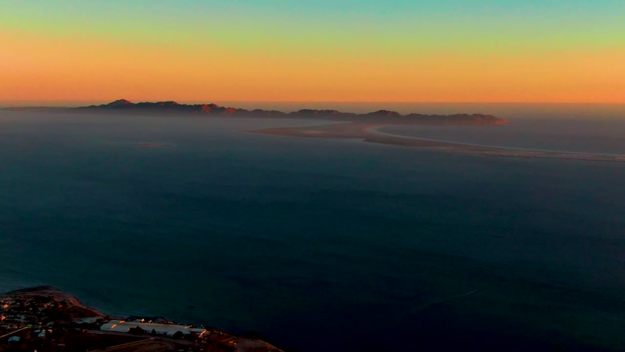Wide aerial view over dark ocean toward fog-covered islands near San Carlos, Baja California Sur, with an orange horizon fading into blue sky and no visible people.
