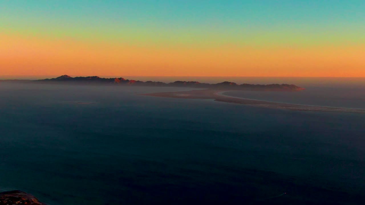 Wide 4K aerial view near San Carlos, Baja California Sur with open ocean in shadow, low mist across distant islands, warm sunset color near the horizon, and no visible people.