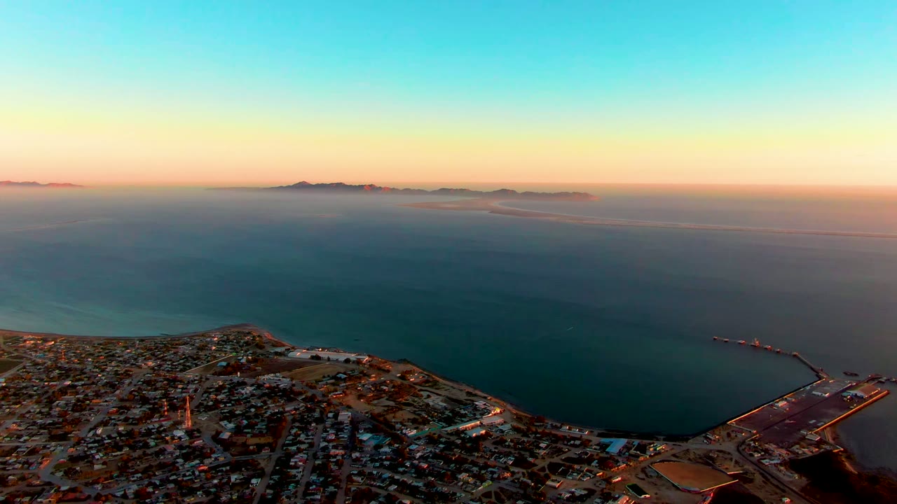 Aerial view of San Carlos, Baja California Sur, with the city and harbor at the bottom, calm sea across the frame, distant islands on the horizon, and no visible people.