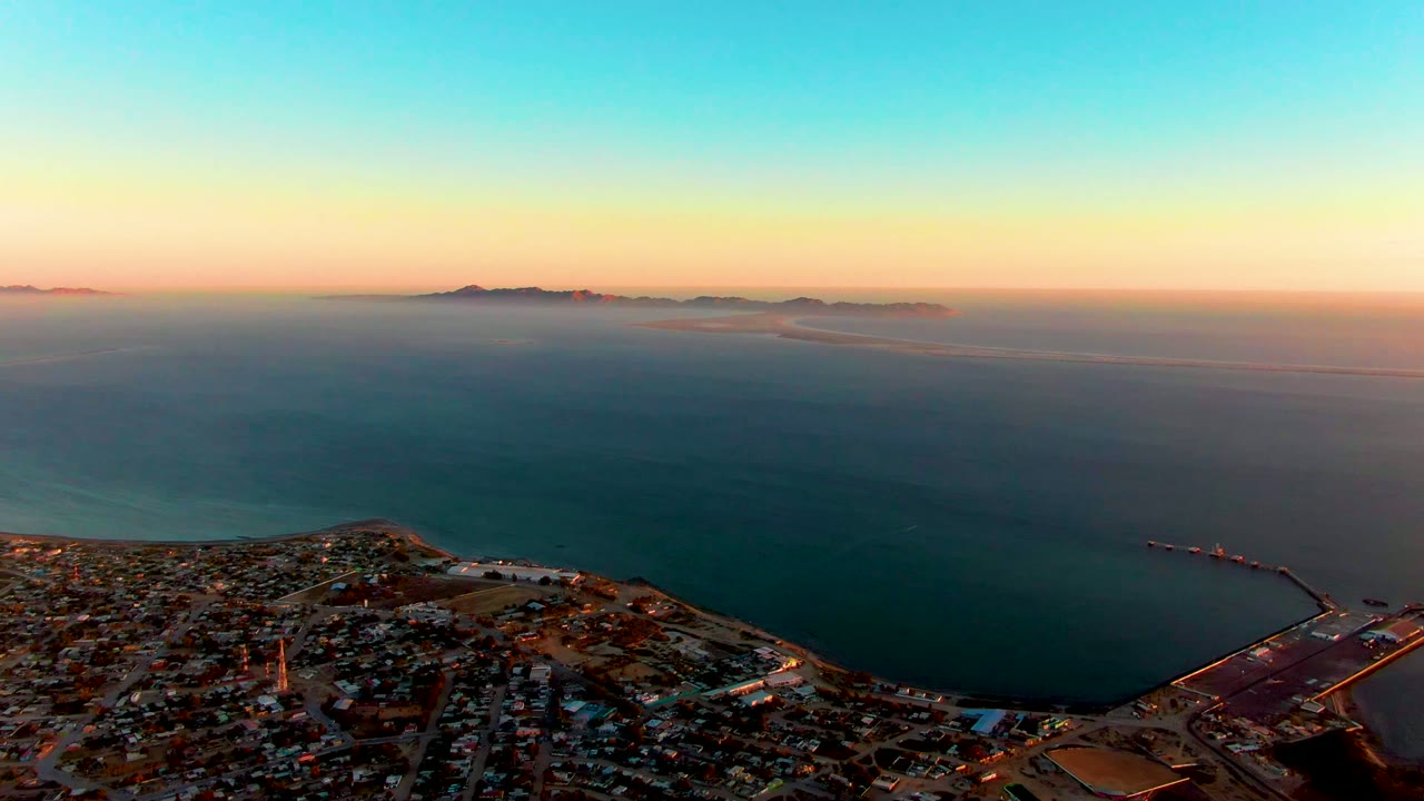 Coastal city and harbor below a wide sunset sea with islands on the horizon.
