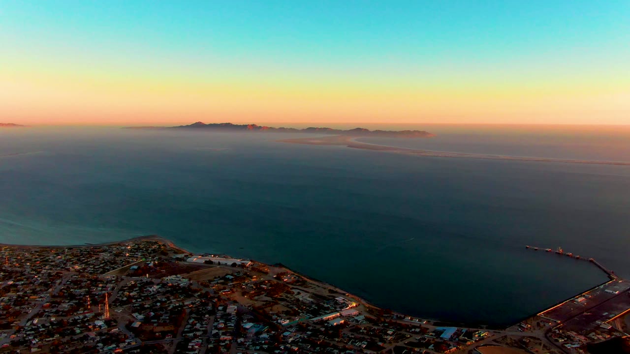 A high aerial frame over San Carlos shows clustered buildings and harbor edges below, open teal water beyond, island silhouettes in the distance, and no visible people.