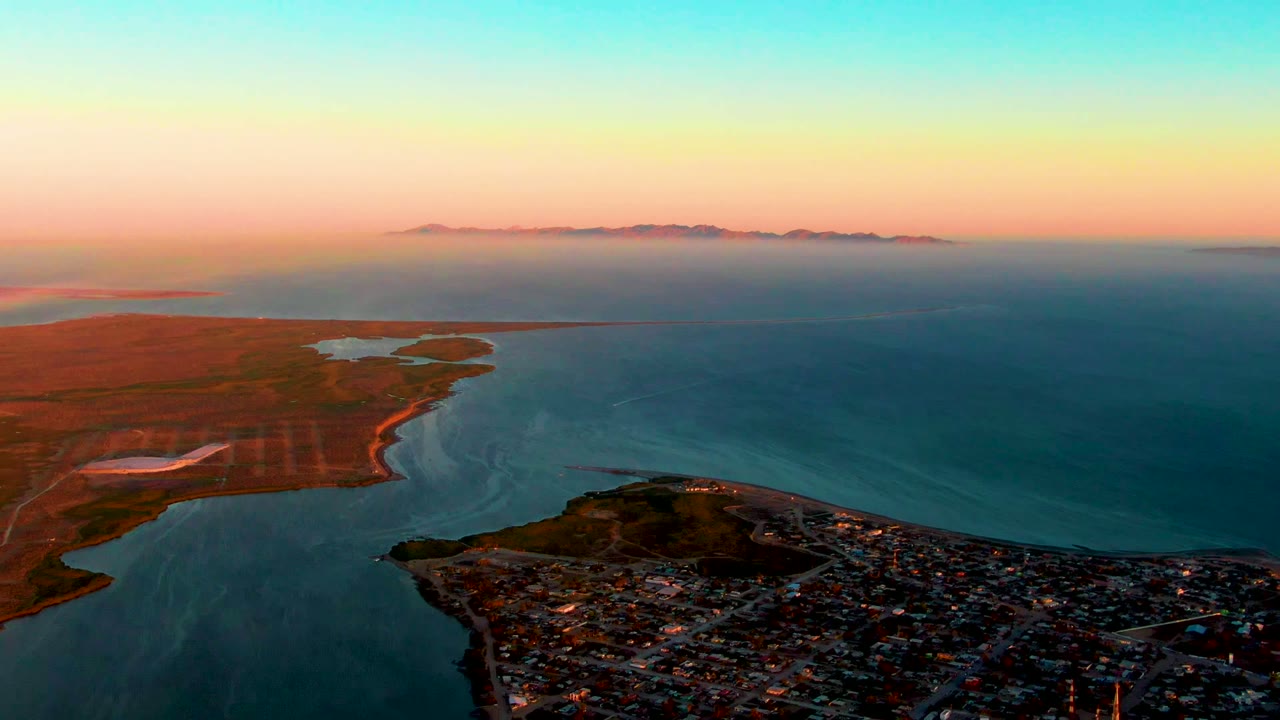 Aerial view over San Carlos, Baja California Sur, with a coastal town in the foreground, blue water wrapping the shoreline, reddish land to the left, distant mountains, and no visible people.
