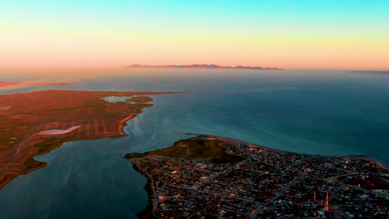 Wide drone view of town, inlet water, reddish coastal flats, and sunset sky.