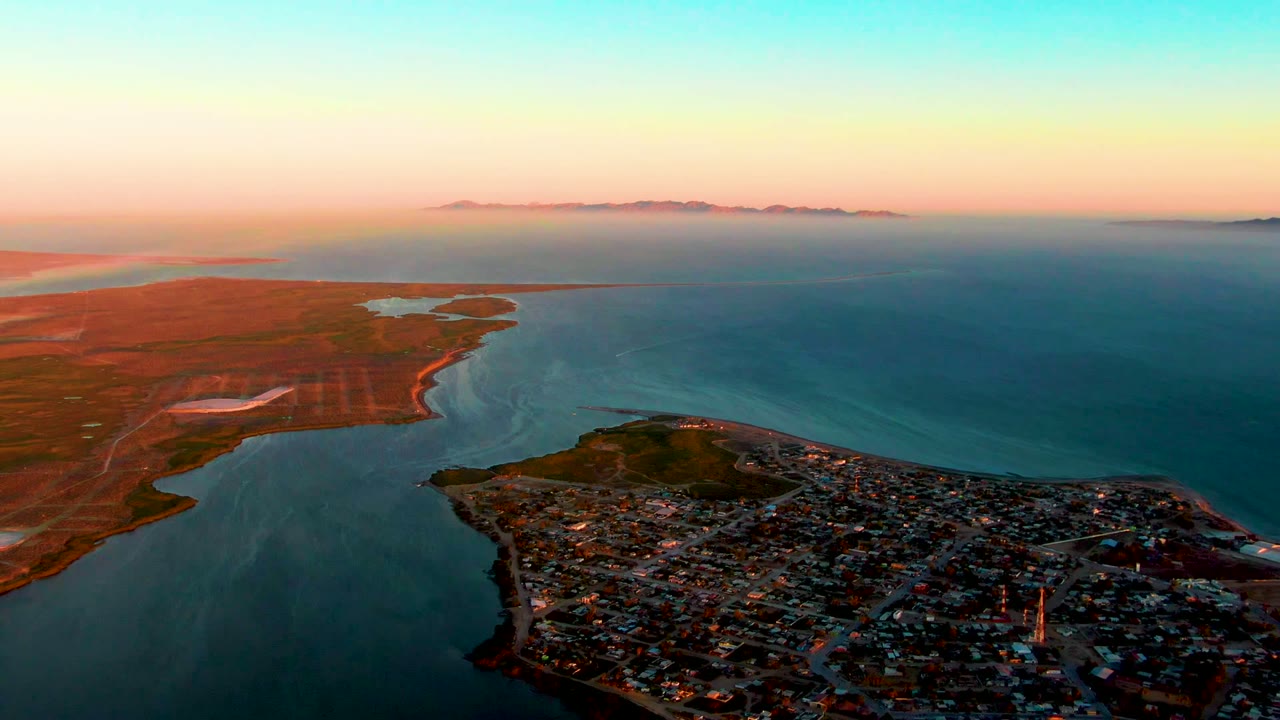 Aerial view of San Carlos, Baja California Sur, with the town spread across a peninsula, open water to the right, reddish land to the left, low mountains on the horizon, and no visible people.
