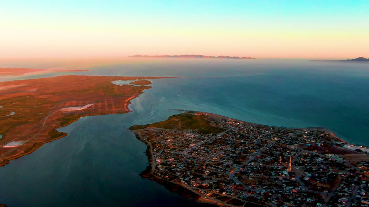 Elevated sunset view of coastal flats, water channel, town edge, and distant horizon.
