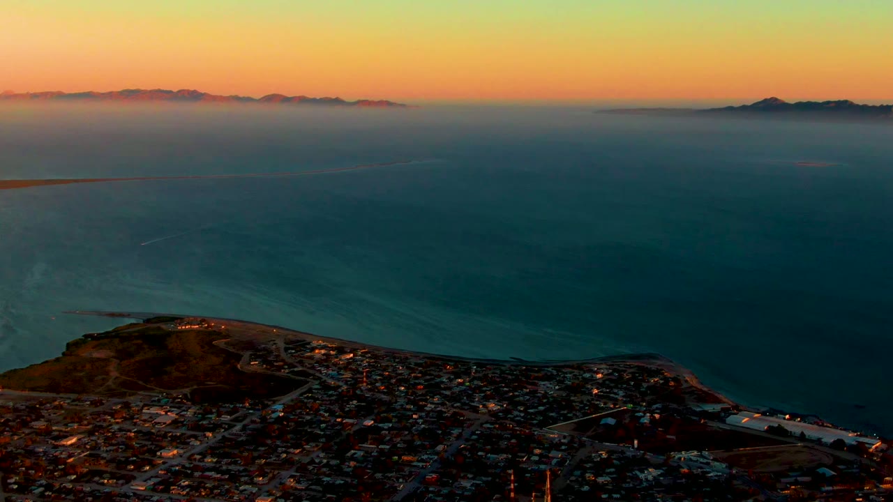 Wide aerial view over San Carlos, Baja California Sur, with the city at the bottom of frame, calm ocean beyond it, distant hills on the horizon, orange sunset light, and no visible people.