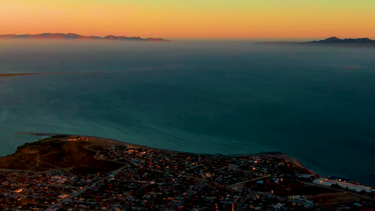 Coastal town lights below a dark ocean and orange-blue sunset horizon.