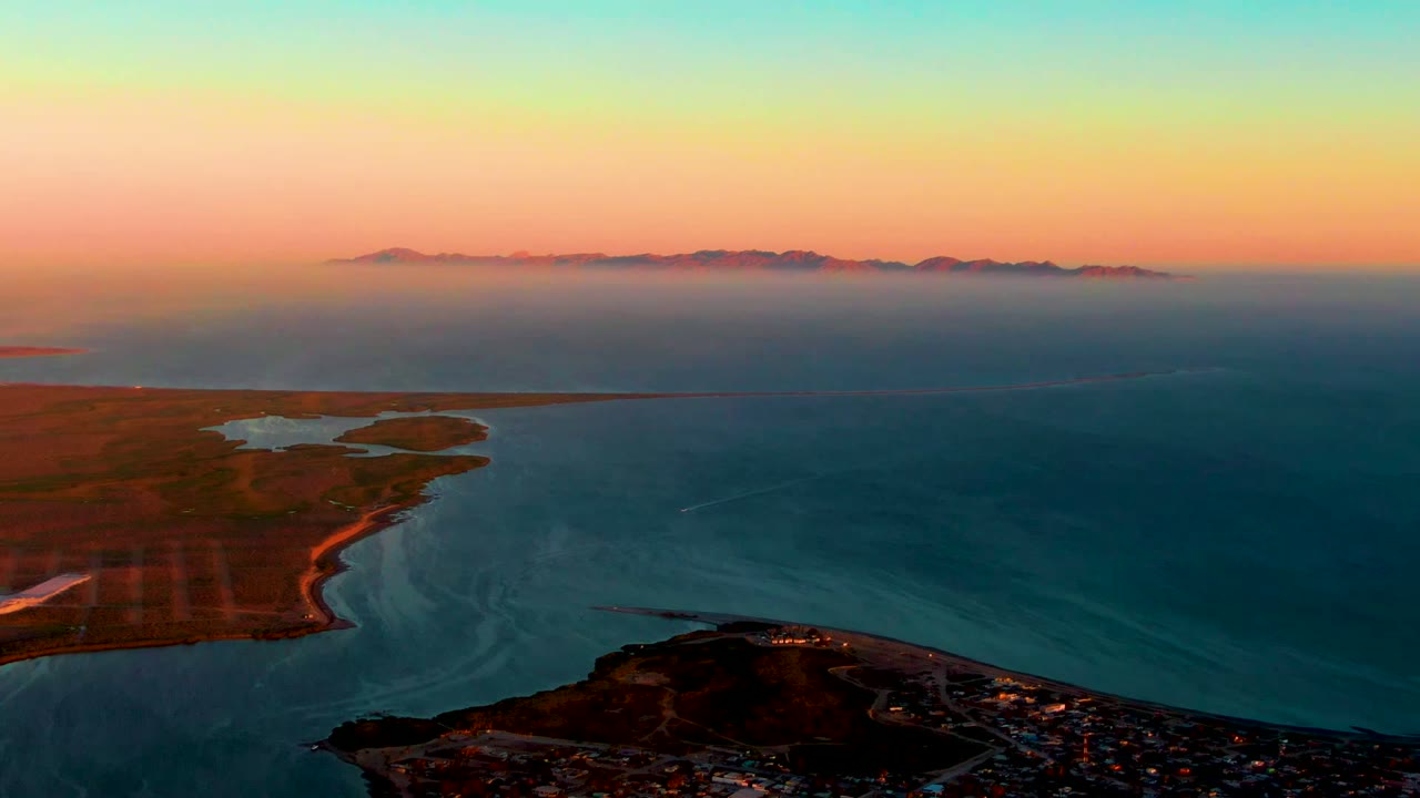 Opening aerial frame over San Carlos, Baja California Sur, with teal ocean currents, a curved shoreline, distant mountains, warm sunset sky, and no visible people.
