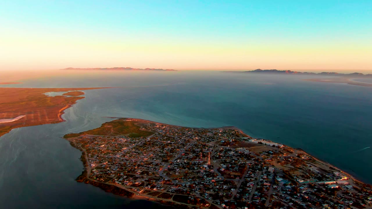 San Carlos, Baja California Sur seen from above at sunset, with the town in the lower frame, blue water around the peninsula, distant islands on the horizon, and no visible people.