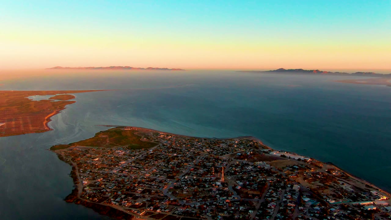San Carlos appears from above with the neighborhood curving along the peninsula, darker blue water to the right, low landforms on the horizon, and no visible people.