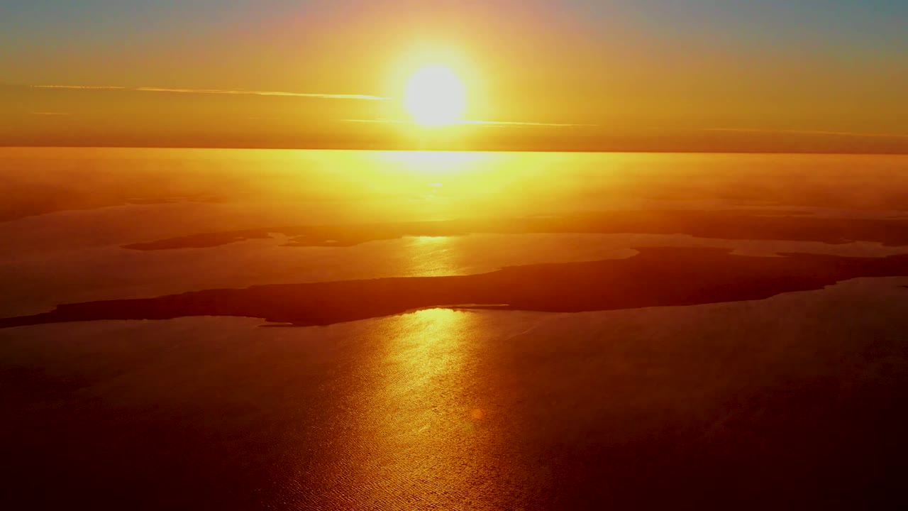 Aerial sunrise view near San Carlos, Baja California Sur, with the sun left of center, orange reflection on the ocean, dark island silhouettes, layered clouds, and no visible people.