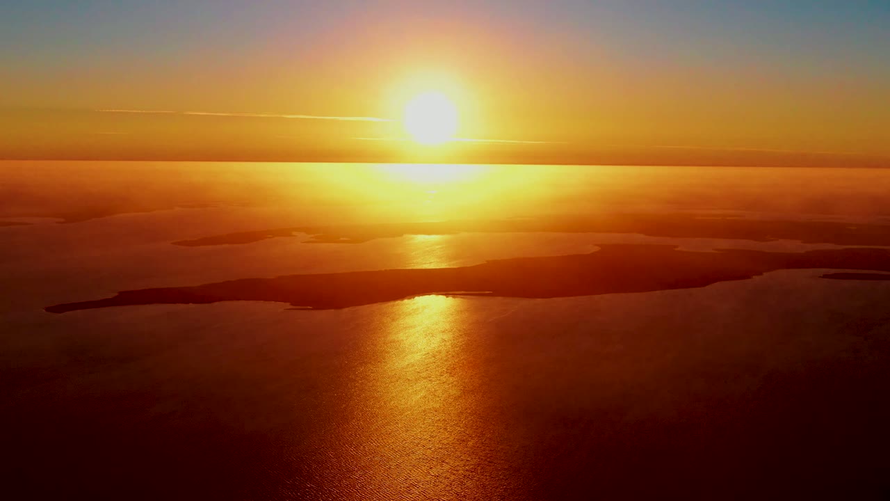 San Carlos, Baja California Sur aerial sunrise with the sun high above the horizon, copper water below, distant island shapes, soft cloud bands, and no visible people.