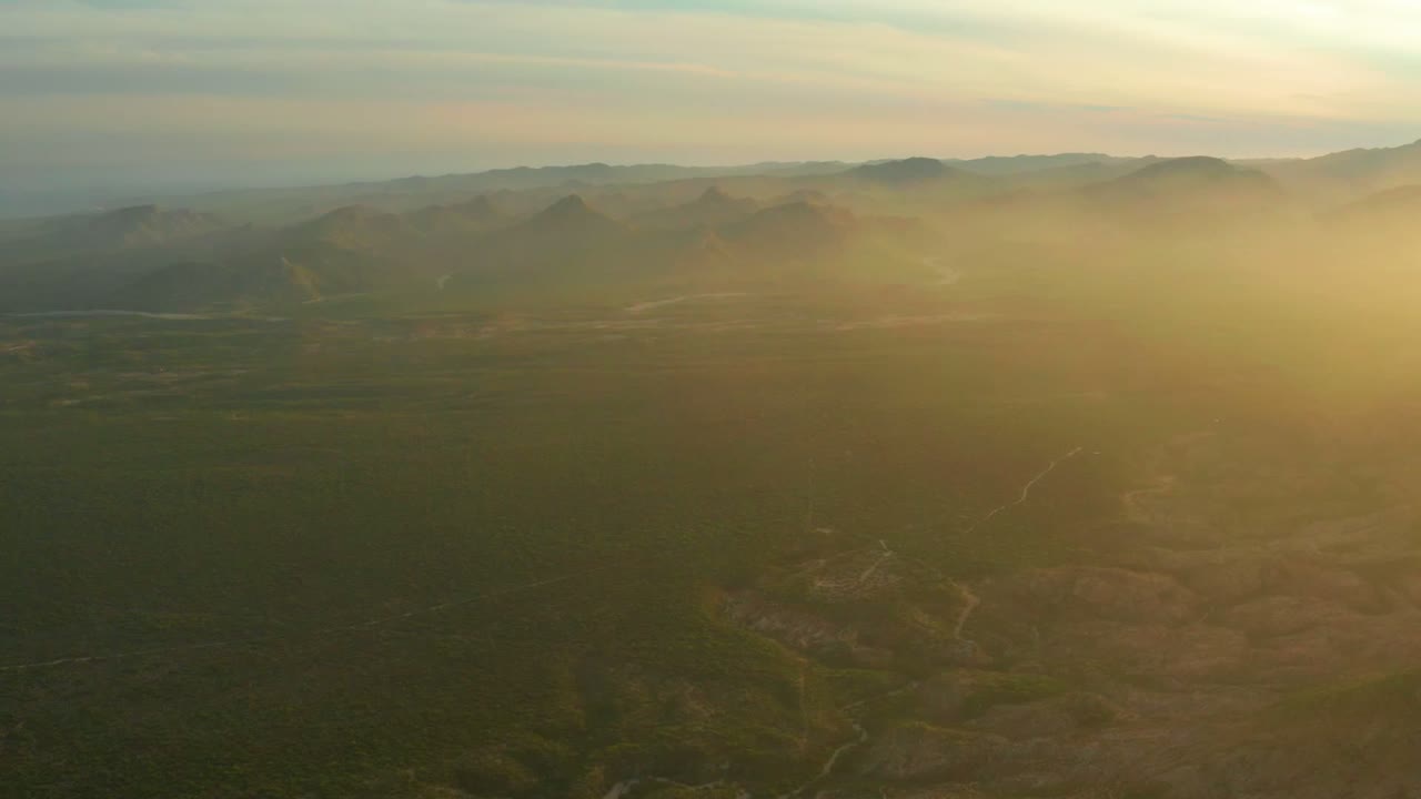 Aerial frame over Cabo Pulmo, Baja California Sur, showing dark green valley terrain, brown open ground, a hazy mountain ridge, warm light from the right, and no visible people.