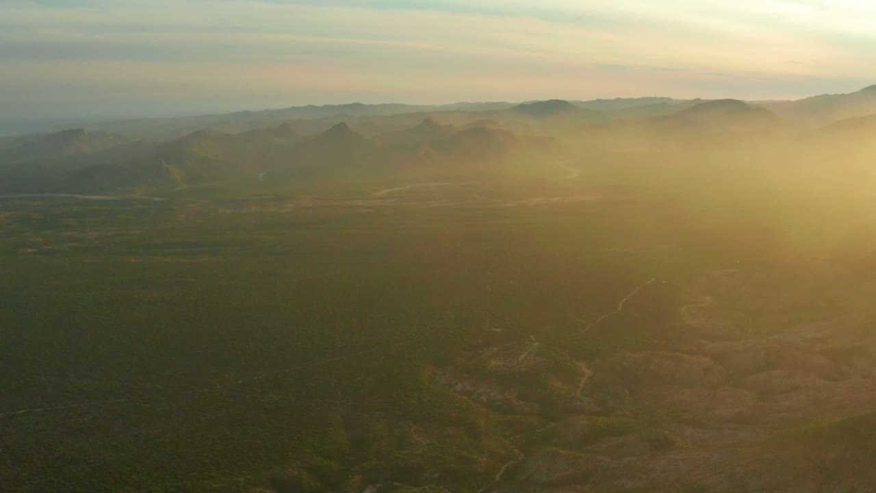 Aerial view in Cabo Pulmo with low mountains in the background, dark valley land in the lower frame, warm sun haze across the right half, and no visible people.