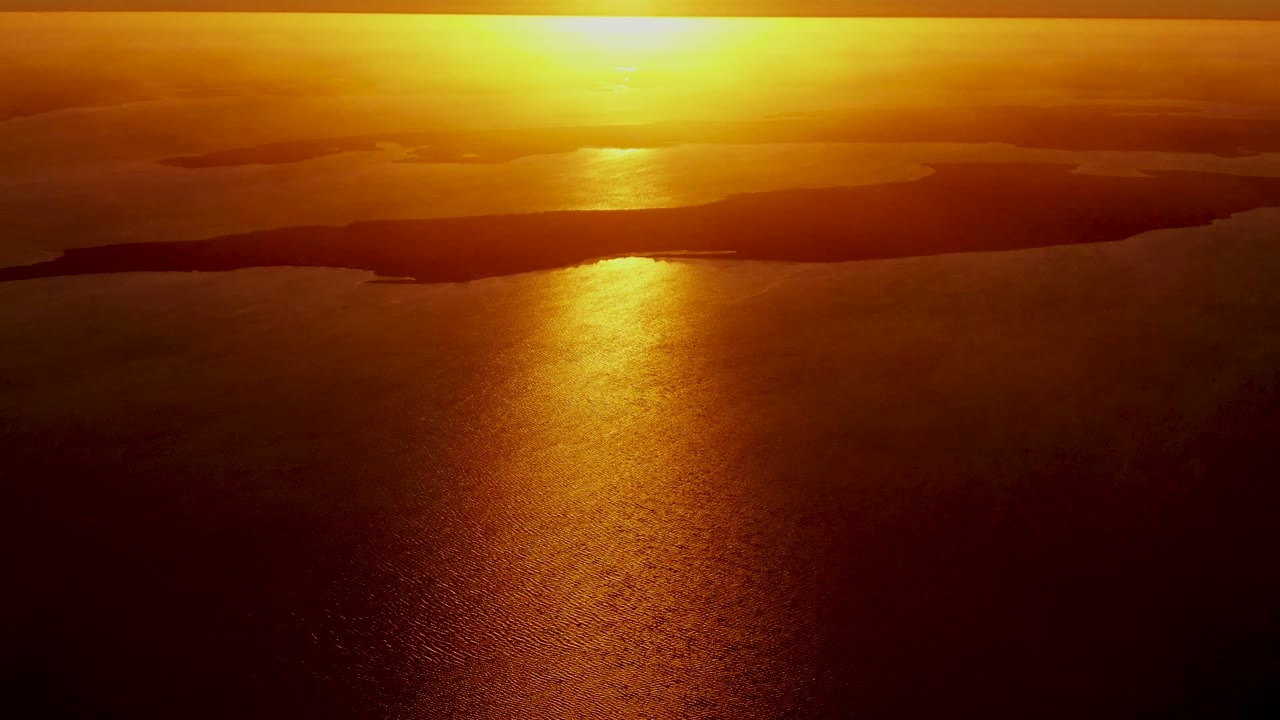 Warm sunset light spreads over calm ocean with dark land on the horizon.