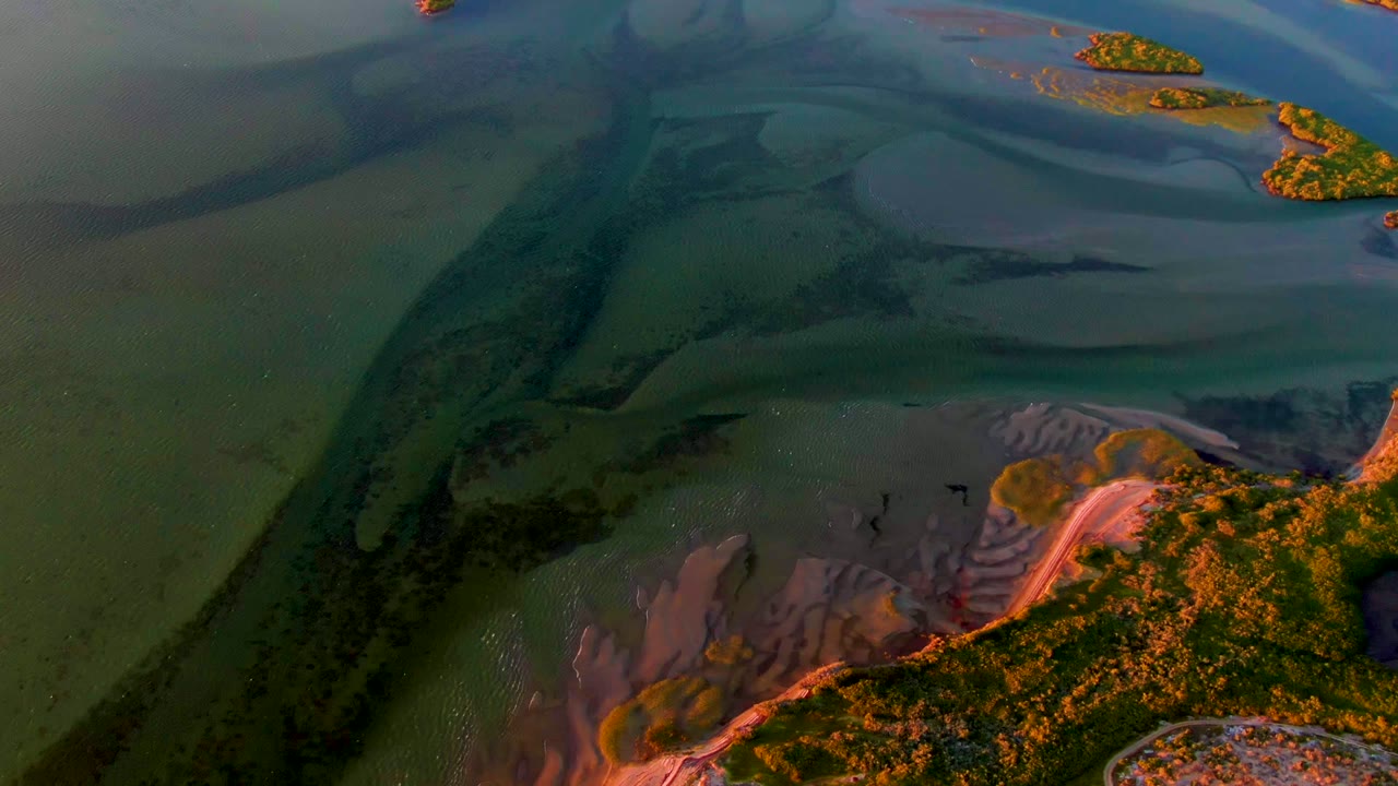 Aerial view over San Carlos, Baja California Sur, showing green water channels, dark sandbars, small islands near the top edge, orange sunset light on the lower shoreline, and no visible people.
