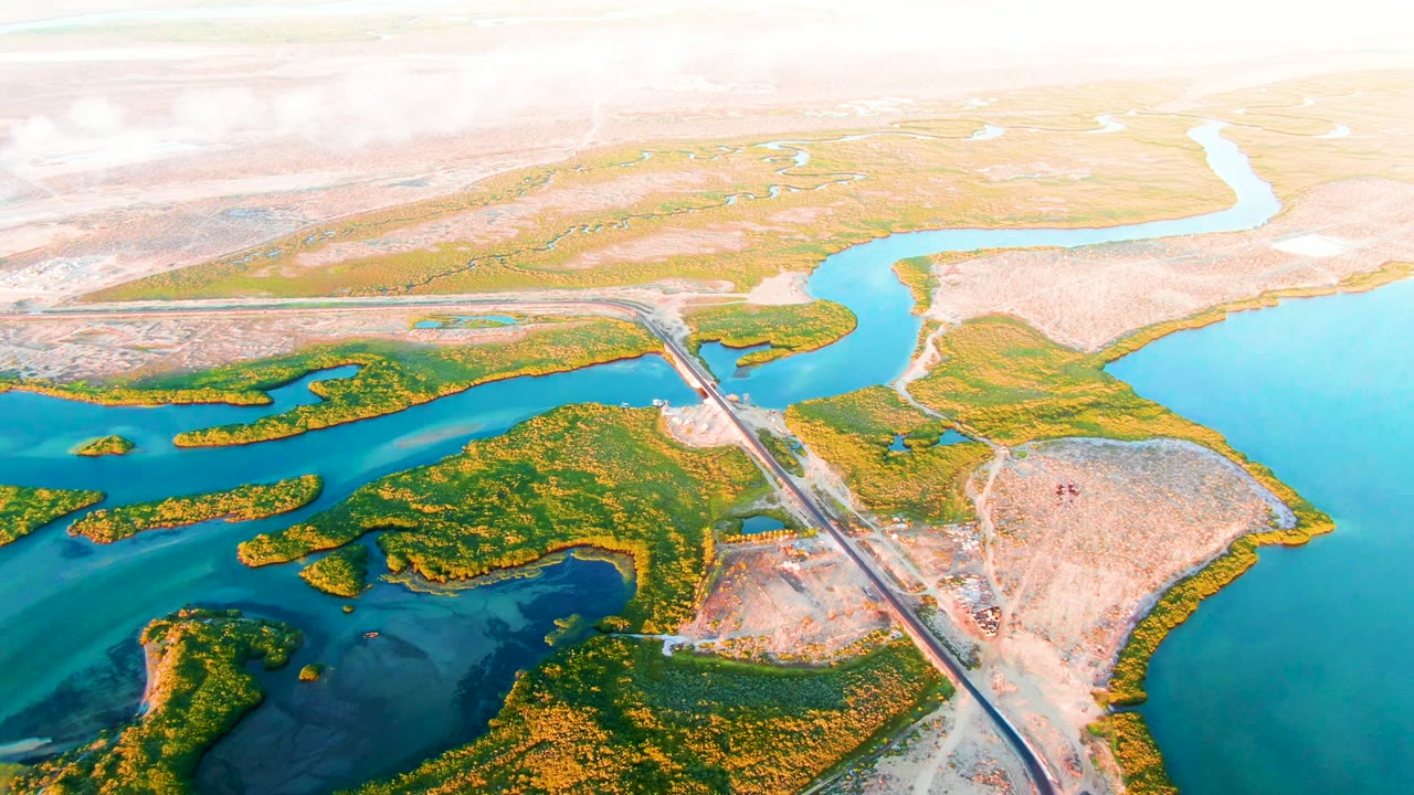 Wide aerial frame over San Carlos, Baja California Sur showing a bridge over blue-green water, vegetated islands, pale dry land, bright haze, and no visible people.