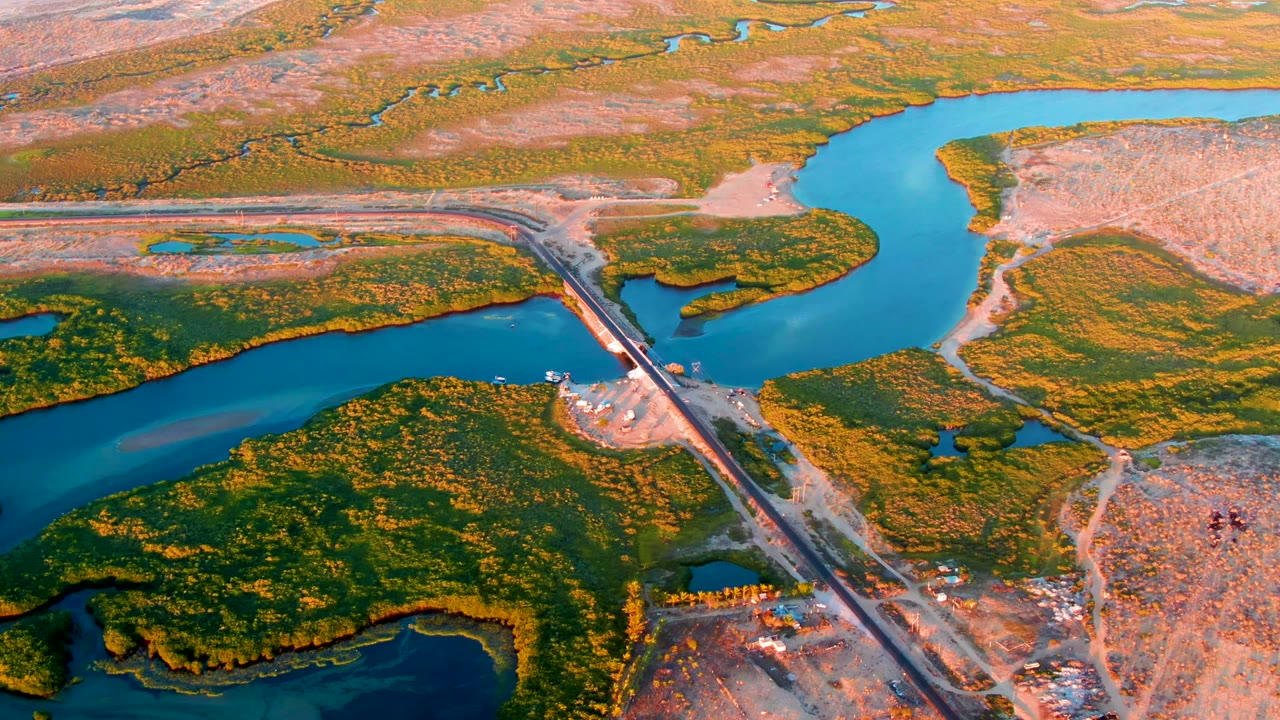 Aerial frame from San Carlos, Baja California Sur, Mexico, showing a straight crossing over a turquoise river, green wetland islands, dry brown flats, and no visible people.