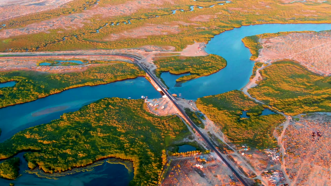 Drone view of a river bend, wetland islands, and a straight crossing near construction land.