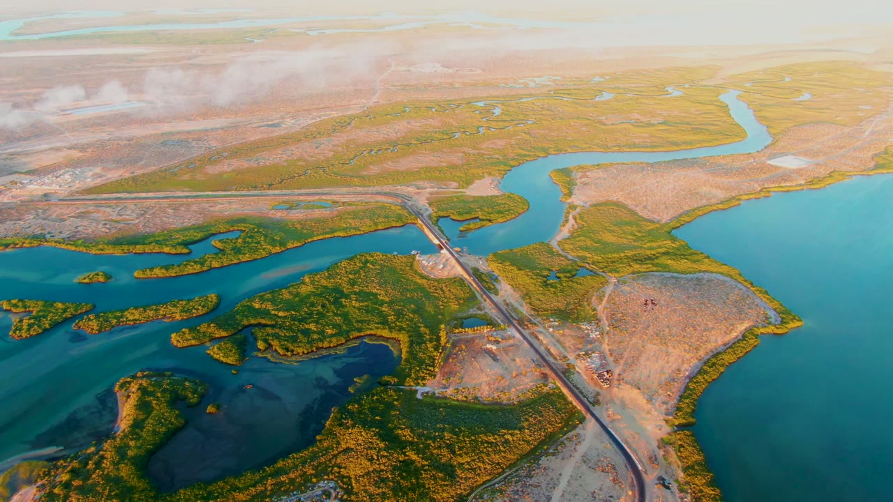 Opening aerial frame over San Carlos, Baja California Sur wetland waterways, with green marsh islands, tan flats, a straight road crossing the water, low cloud in the distance, and no visible people.