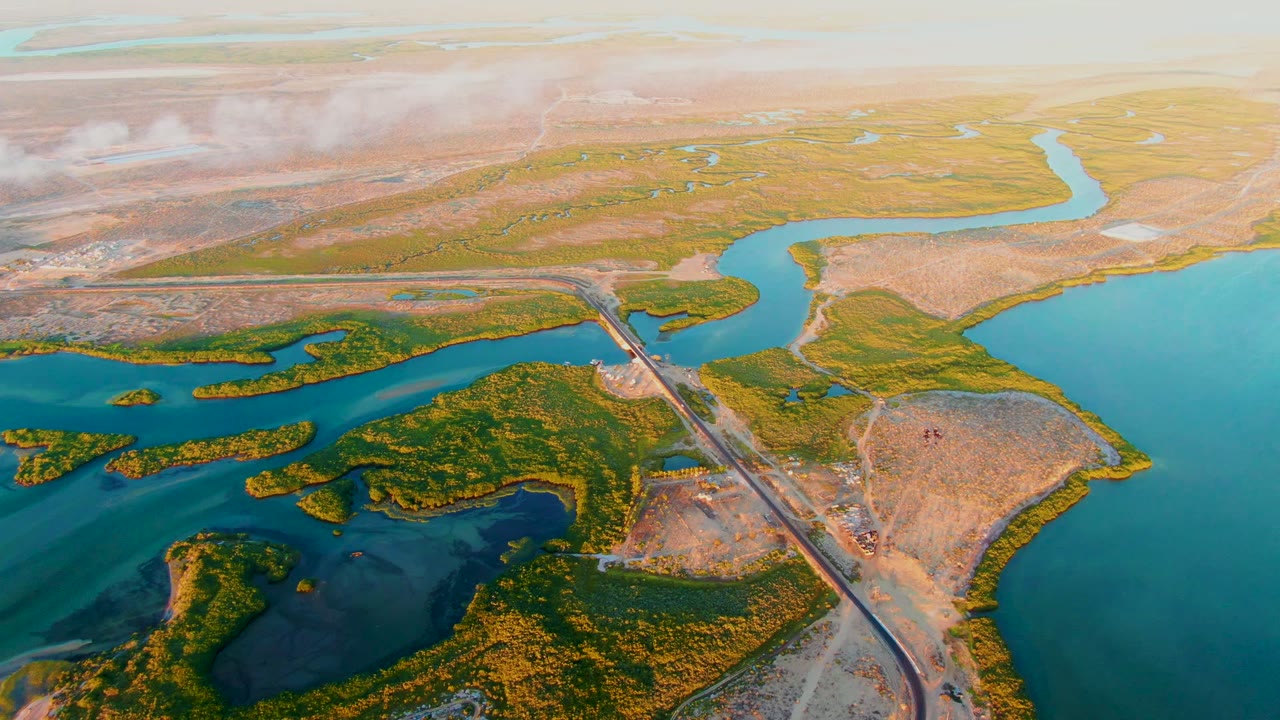 Drone view of marsh islands, blue water channels, tan land, road crossing, and pale cloud.