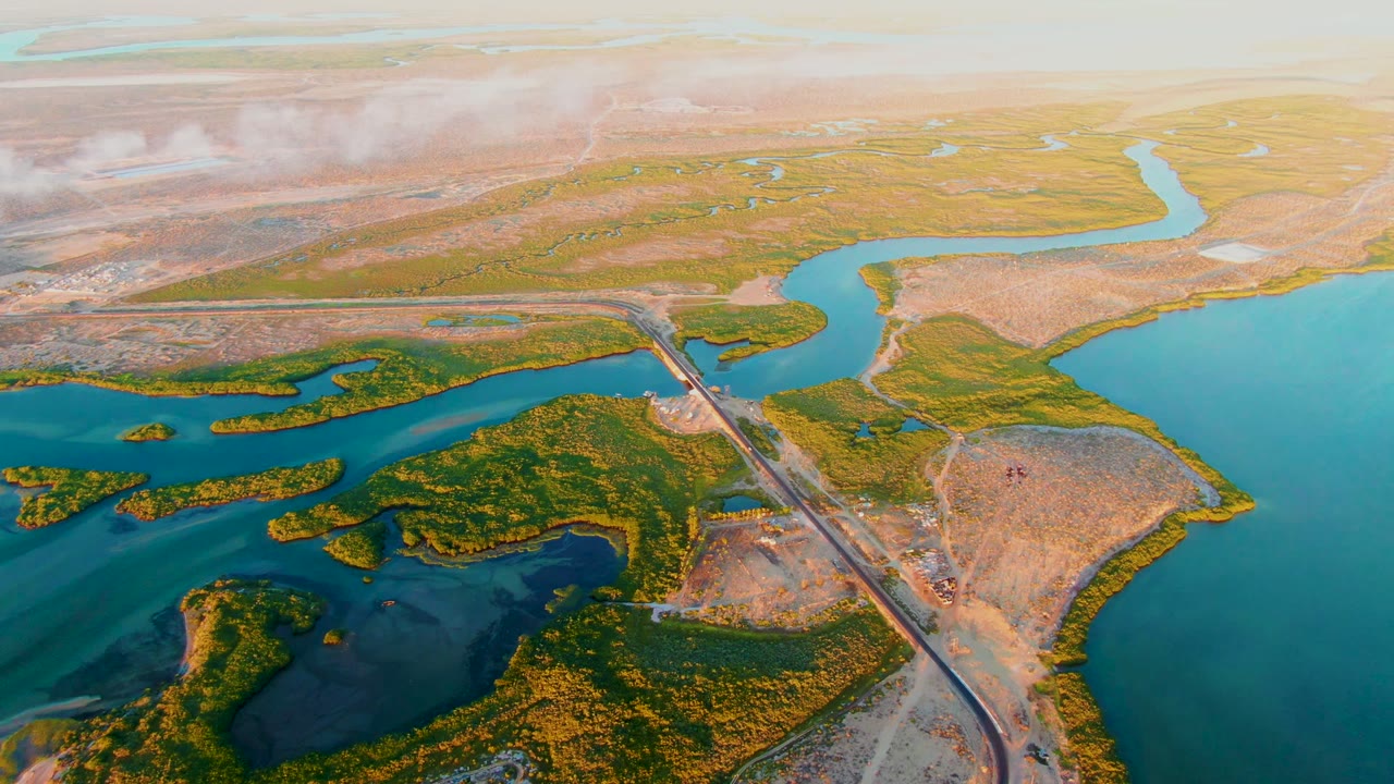At 0:02, the San Carlos wetland fills the frame with turquoise water on the right, darker channels on the left, a diagonal road crossing, hazy clouds above, and no visible people.