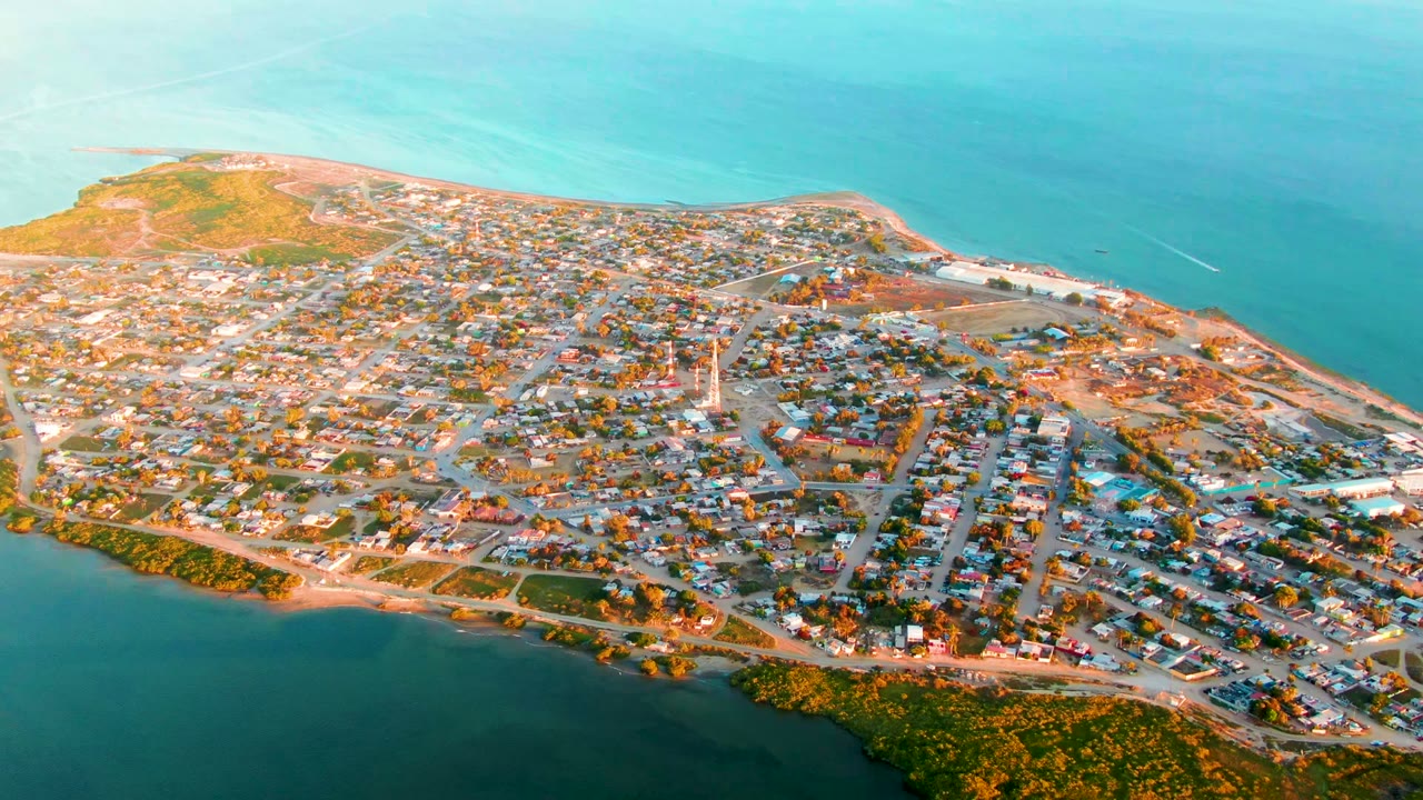 High aerial view over San Carlos, Baja California Sur, showing dense low buildings, roads, shoreline vegetation, and blue water on both sides with no visible people.