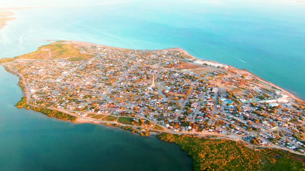 High drone view of San Carlos, Baja California Sur, with a compact settlement below, turquoise sea across the horizon area, a narrow shoreline edge, and no visible people.