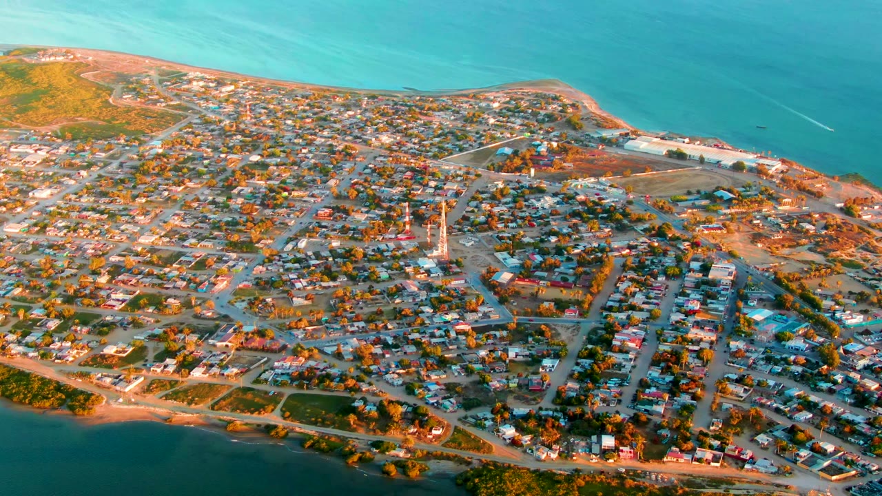 Aerial frame over San Carlos, Baja California Sur showing compact residential streets, low houses, green shoreline, sandy beach, turquoise ocean, and no visible people.