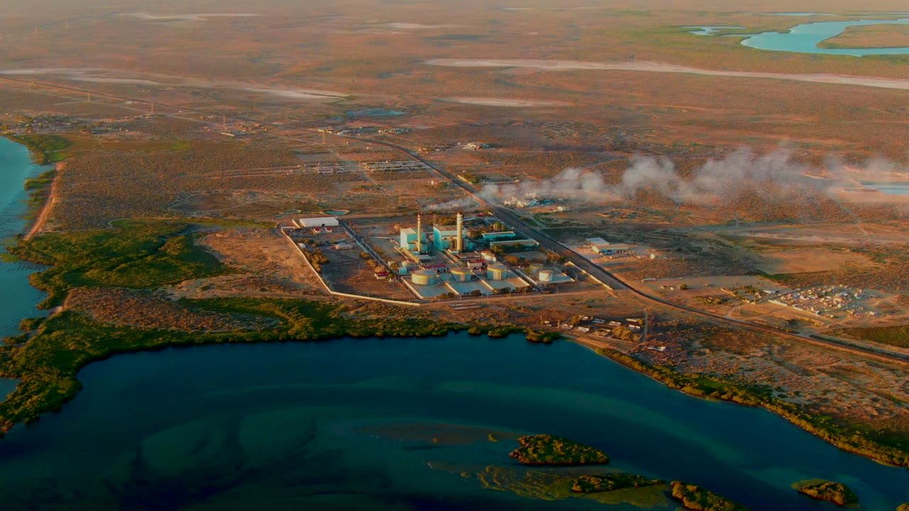 Opening frame over San Carlos, Baja California Sur, showing an industrial plant beside blue-green water with a white smoke plume drifting across dry terrain and no visible people.