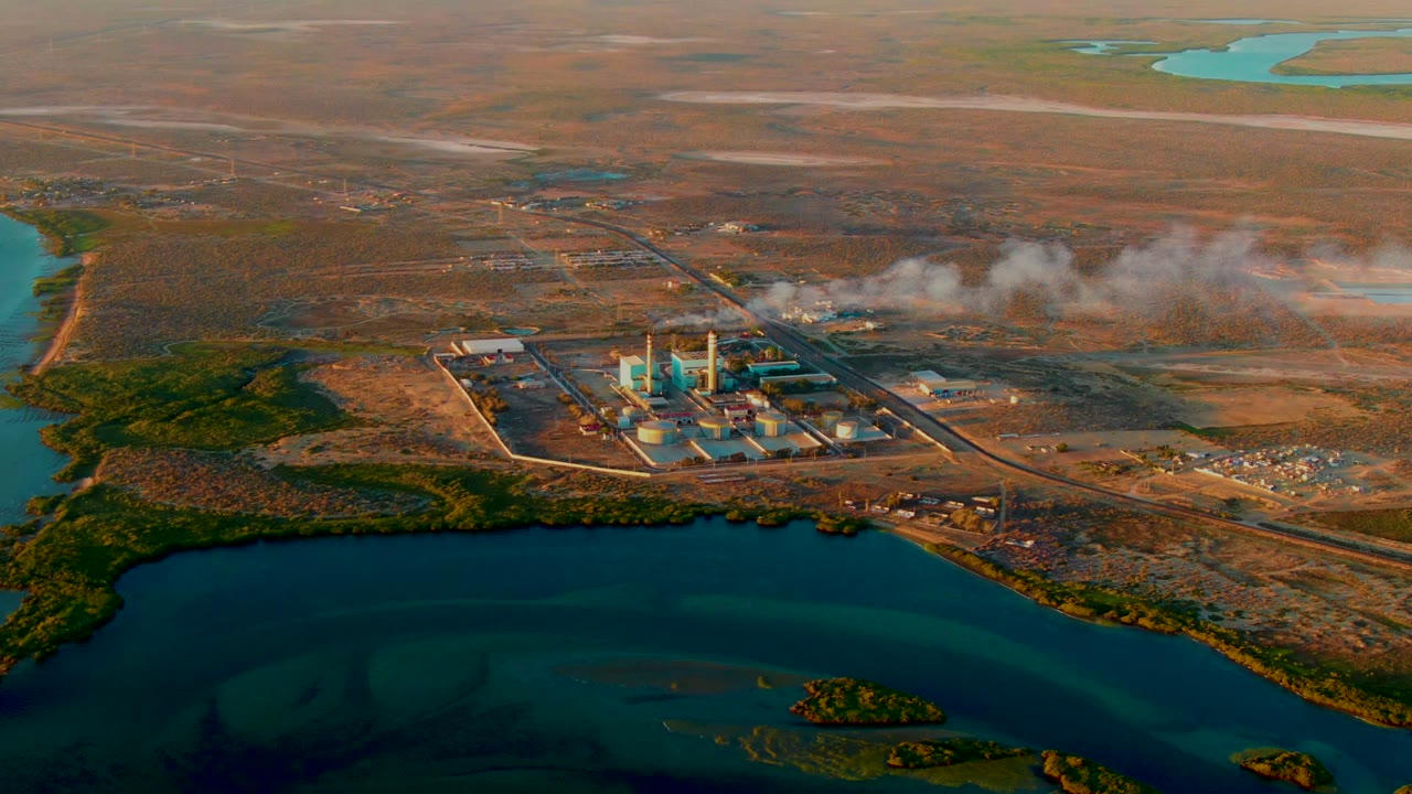 Aerial industrial complex beside water with smoke over a dry landscape.