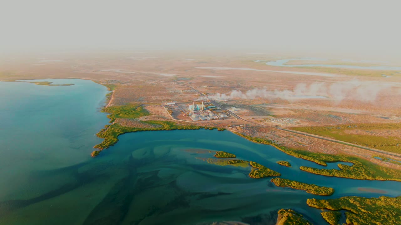 Opening aerial frame over San Carlos, Baja California Sur, with blue-green water, small green islands, a shoreline industrial plant, pale smoke, dry brown land, and no visible people.