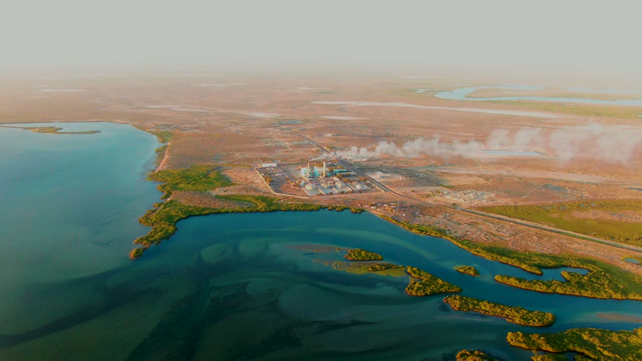 Wide drone view of smokestacks and smoke near a large body of water and dry terrain.