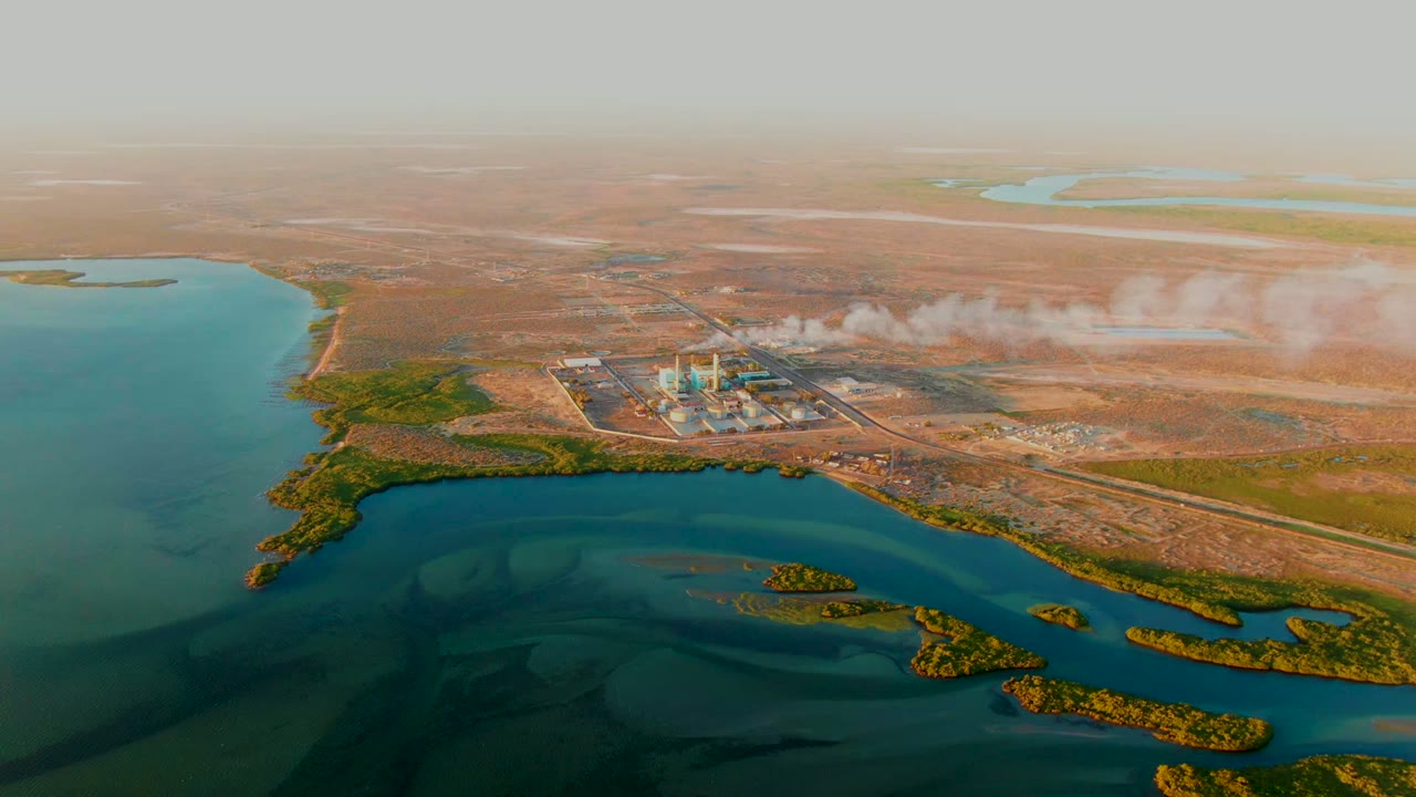 A wide aerial frame over San Carlos, Baja California Sur, with turquoise water filling the left foreground, the plant near the center, smoke trailing across dry land, and no visible people.