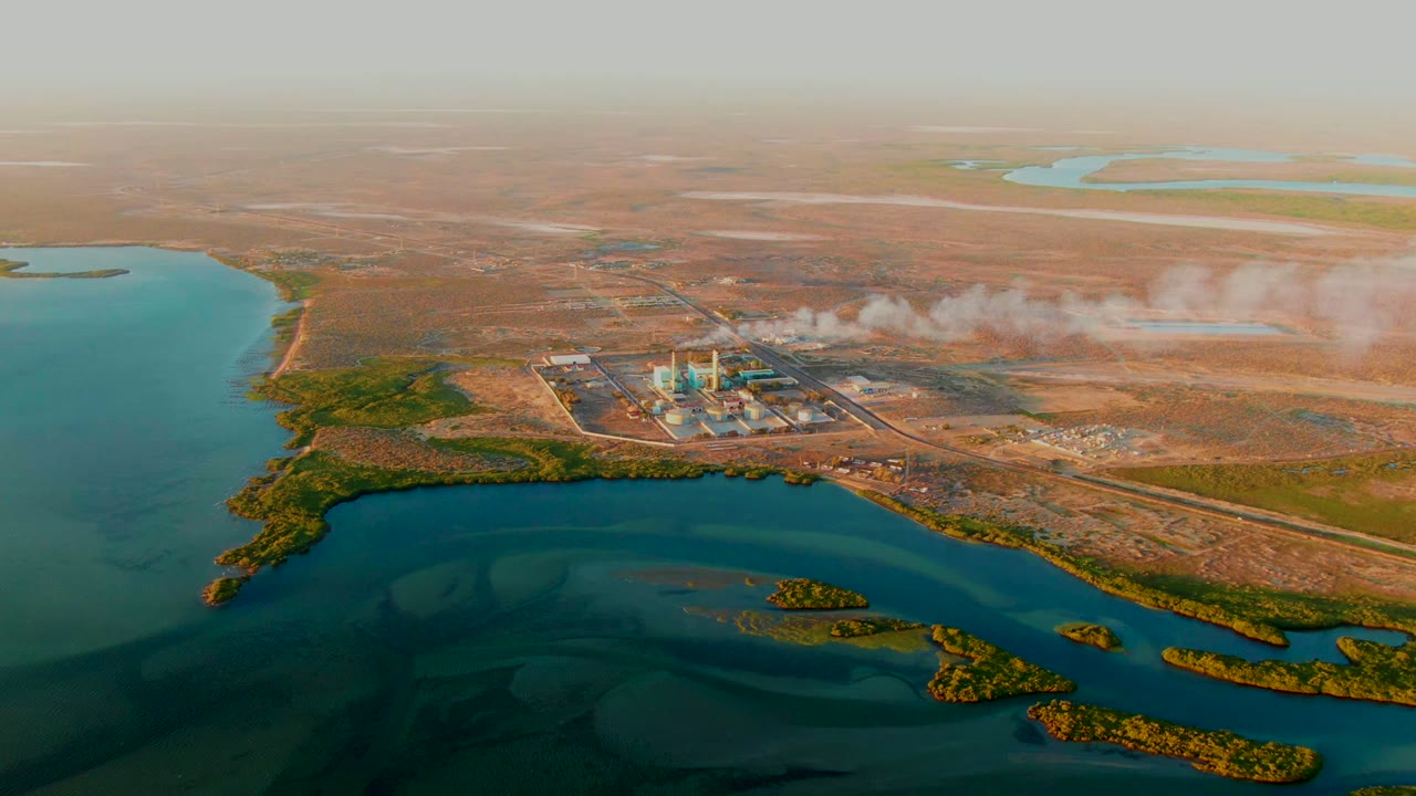 Drone perspective of an industrial plant emitting smoke beside dark blue water and arid land.